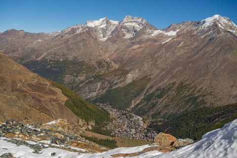 View down the valley to a village surrounded by snow covered mountains under a bright blue sky,