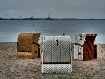 Three beach chairs stand on the sandy beach in front of a calm sea under a cloudy sky. Ships are