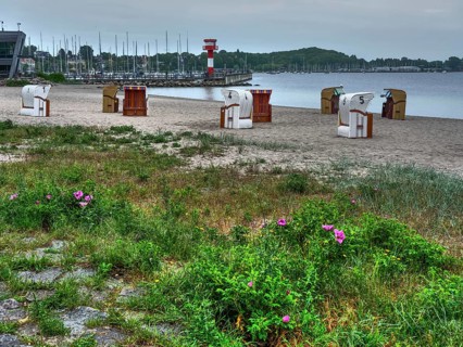 Scattered beach chairs on a sandy beach with a harbour and sailing boats in the background. The sky