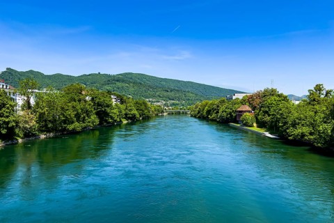 A calm river flows through a landscape of green trees and hills under a blue sky, Olten, Solothurn,