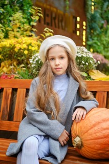 Cute little girl in a beret and woolen coat sits on bench leaning on pumpkin