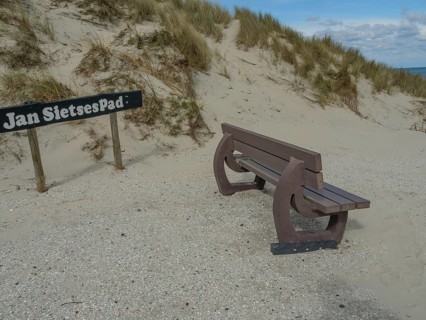 A bench by the sand dunes, next to it a sign with the inscription 'Jan Sietsepad', on it a partly