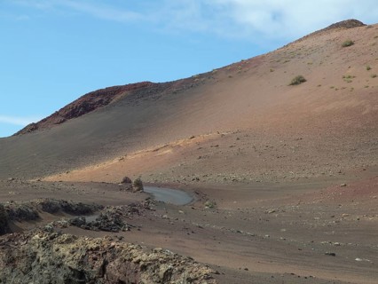 A rocky volcano with a narrow road, surrounded by lava and ash under a slightly cloudy sky, barren