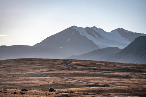 Gravel road through autumnal plateau with brown grass, glaciated and snow-covered peaks, Ak Shyrak