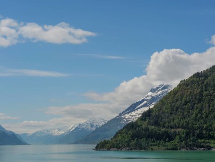 Snow-capped mountains next to a calm lake under a clear blue sky with some clouds, greenish