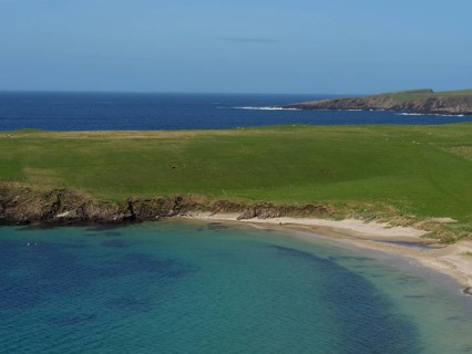 Green grassy landscape along a calm bay with clear blue water under a wide sky, golden beach with
