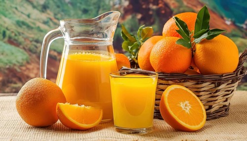 A glass and a jug of orange juice next to a basket of oranges and green leaves in a natural setting