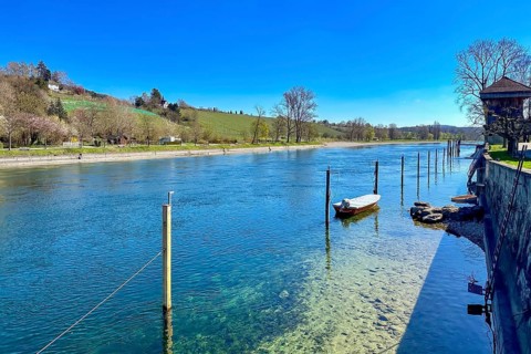 Clear river landscape with calm water, boat moorings and green hills in the background in bright