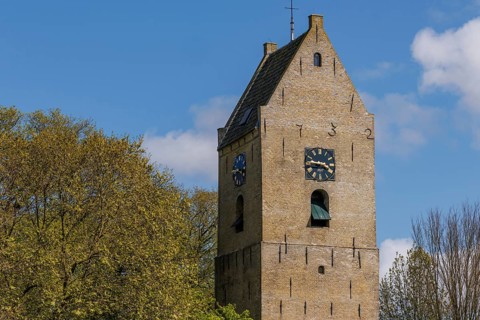 A medieval bell tower with clocks on both sides against a blue sky, historic houses and a church