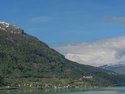 Sunny landscape with snow-covered mountains and houses on the lakeshore under a blue sky, greenish