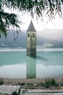 Sunken tower in Lake Reschen with mountains in the background and a gravel path in the foreground.