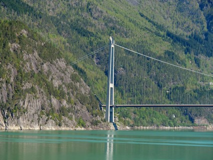Imposing bridge over bright green water, framed by steep, forested mountains under a blue sky,