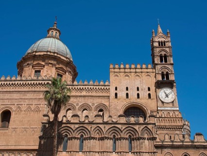 Historic cathedral with striking dome, clock and detailed towers, under a bright blue sky, two red