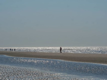 Single person walking along a wide beach, other people visible in the distance, glittering sea