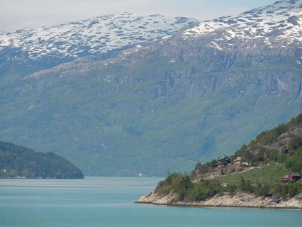 A calm lakeshore with mountains and wooded hills, under a clear blue sky with clouds, greenish
