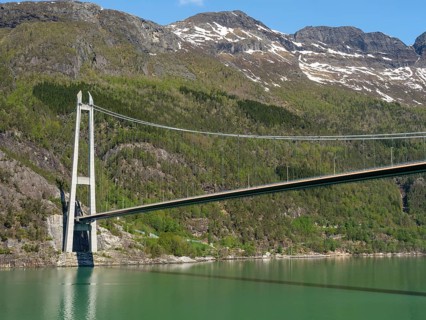 Large bridge over calm water, framed by forested mountains and rocks under a bright blue sky,