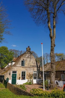 An old farmhouse with flagpole and blue door, surrounded by tall trees and brick houses, historic