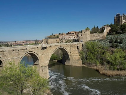 Stone bridge over a river with historic buildings and vegetation under blue sky, historic stone