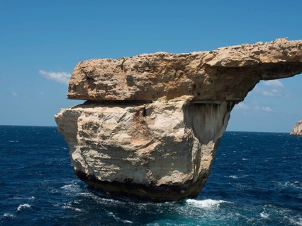 Massive rock arch towers over the sea, surrounded by cliffs under clear blue skies, many historic