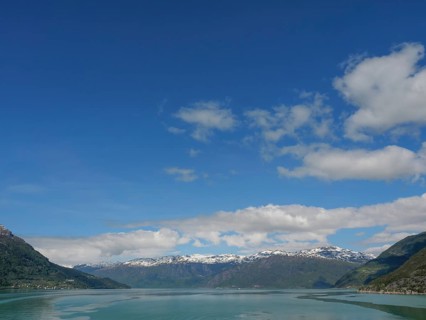 Expansive view of a mountain range under a clear blue sky with some clouds and a calm lake,