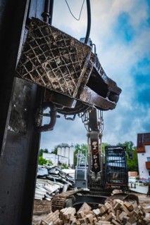 Close-up of the grab of an excavator in the foreground, working surrounded by rubble under a cloudy