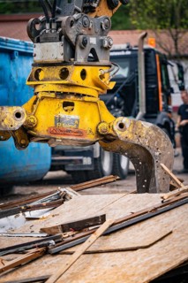 An excavator with a hydraulic grab stacks wood debris while a worker stands nearby, demolition,