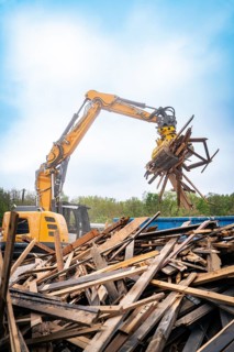 Large excavator lifts wood debris on a construction site under a cloudy sky and numerous