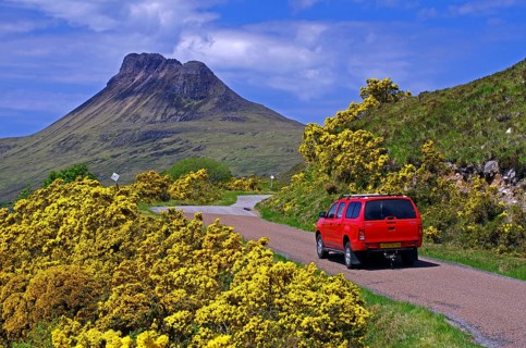 Car driving on a road framed by intensely flowering gorse, high barren mountains, spring, Ullapool,