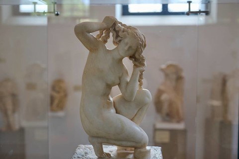 Aphrodite, crouching, Kneeling marble sculpture of a woman braiding her hair, surrounded by other