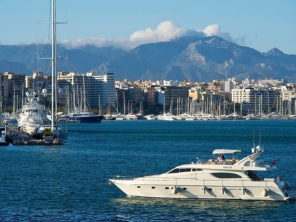 Harbour with a yacht in the foreground, city and mountains in the background under a cloudy blue