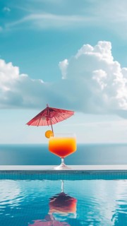 A glass of orange juice with a red umbrella on top of it placed at the edge of a pool, near the
