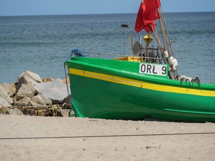 A green fishing boat with a red flag lies on the beach near rocks, green and yellow fishing boats
