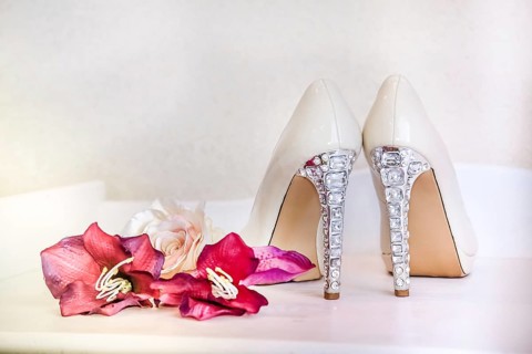 Fancy white high heels with rhinestone details next to pink flowers on a white bench creating an
