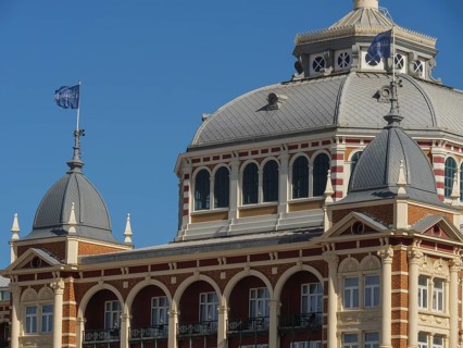 Historic building with domes and flags under a blue sky, parasols and a pier on the beach of