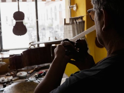 A luthier seen from behind works on a violin near a well-lit window, tools spread out on the bench