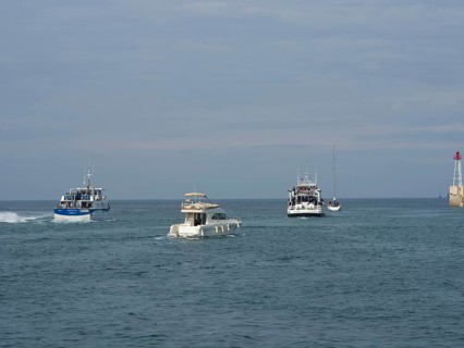 Boats sailing on the sea towards a harbour, accompanied by the wide sky and a lighthouse in the