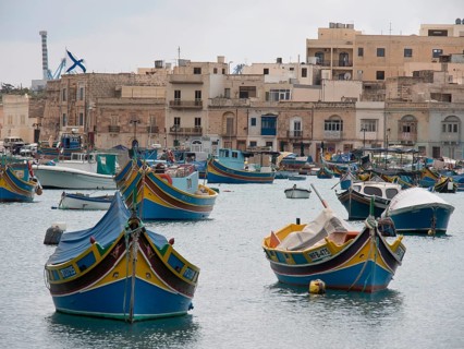 Several colourful boats in the harbour, behind them a town with many residential buildings and calm