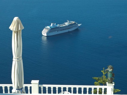 A cruise ship in the blue sea near a coastline with a parasol and railing in the foreground, The