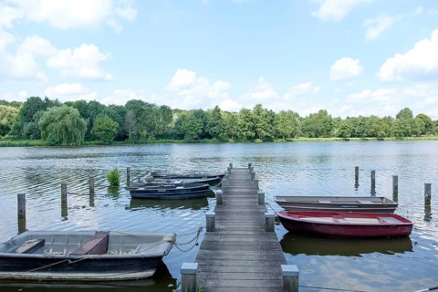 A wooden jetty leads onto a lake with boats, surrounded by trees and a cloudy sky, small lake with