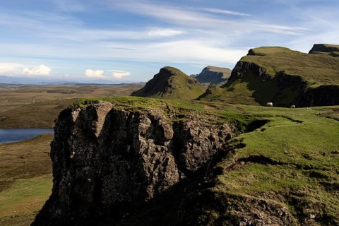 Dramatic cliffs and green slopes in an expansive mountain landscape under a blue sky with scattered