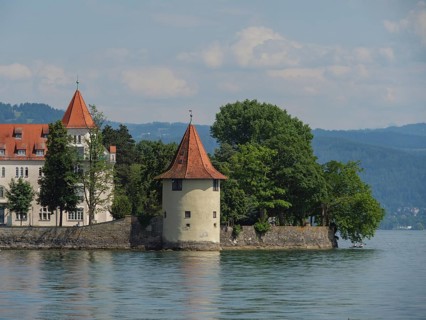 A tower with a red roof and a building on the shore of a lake, surrounded by trees and mountains in