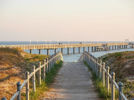 A long wooden walkway leads into the sea, framed by grass and promenade lighting in the soft