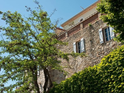 A light-coloured stone house with green trees and shutters under a blue sky, with climbing plants