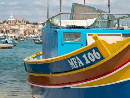 A blue and yellow fishing boat at a harbour with a coastal town in the background, many colourful