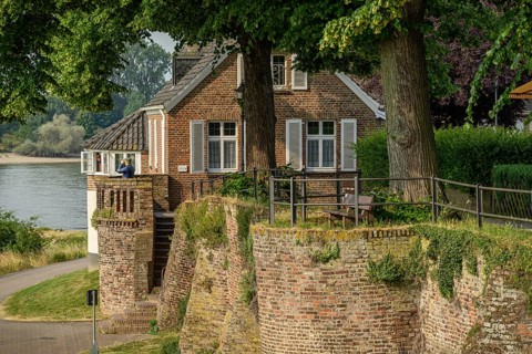 An old house on a riverbank, surrounded by a wall and trees, summery historical landscape, the