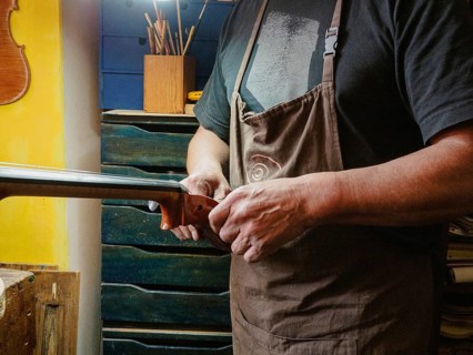 A luthier carefully works on a violoncello head and curl while wearing an apron, surrounded by