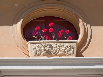 An oval window with a decorative flower box full of red flowers in front of it, the town of mdina