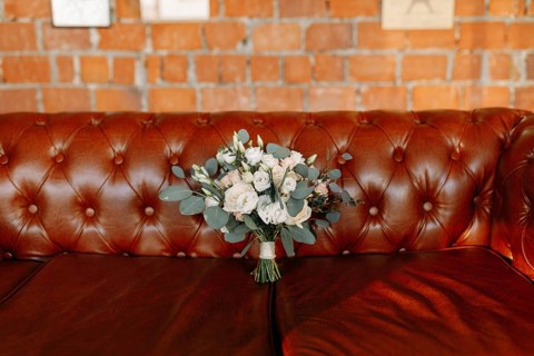 A bouquet of white flowers is placed on a brown leather couch against a brick wall
