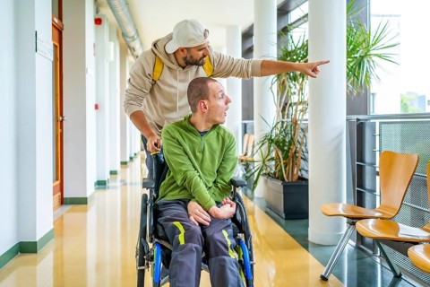 Caucasian young man showing something outside the university to a disabled friend sitting on