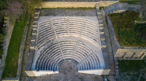 Aerial view of an ancient amphitheatre with stone seating steps surrounded by green areas,